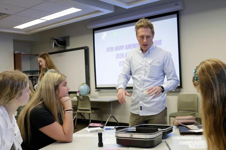 Nate Scholten as a graduate student instructor talking to a table of his students and teaching his course. He is wearing a light blue shirt, and behind him is the projector from the classroom.